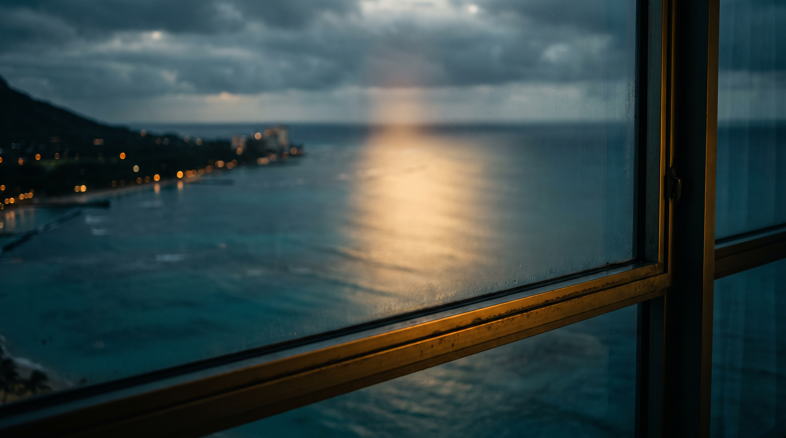 Waikiki hotel window at dawn — a gold light shaft on the Pacific, Diamond Head at left, city lights along the shore, rain on the glass.